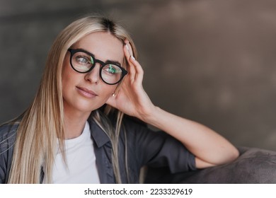 Close up portrait of gorgeous blonde Swedish woman in glasses, grey shirt with long loose hair leaning on hand looks atside toothy smiles, satisfied by career of psychologist. Model posing indoors. - Shutterstock ID 2233329619