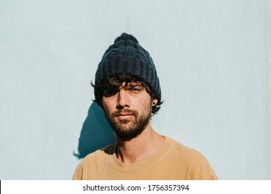 A close up portrait of a green eyes young male with beanie, beard and t-shirt. The handsome male model is looking at the camera. Complementary colors. Light blue background. Copy space