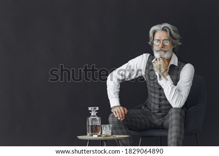 Close up portrait of grinning old-fashioned man. Elegant man in a studio. Gentelman sitting on a chair. Grandfather with a glass of whiskey.