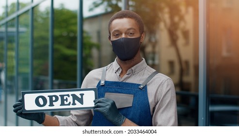 Close Up Portrait Of Handsome African American Young Male Owner In Apron And Mask Standing Outdoor On Street With Open Sign And Looking At Camera. Restaurant Business Coffee House Reopening Quarantine