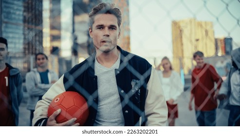 Close Up Portrait of a Handsome Caucasian Man with Red Soccer Ball Posing and Looking at Camera while Standing Behind a Fence on a Rooftop Urban Parking Lot. Diverse Friends Standing Behind Him - Powered by Shutterstock