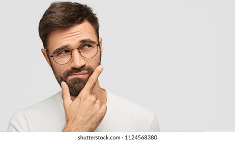 Close up portrait of handsome dark haired male holds chin and looks with hesitsation aside, chooses what to buy between two things, isolaed over white background with copy space for your text