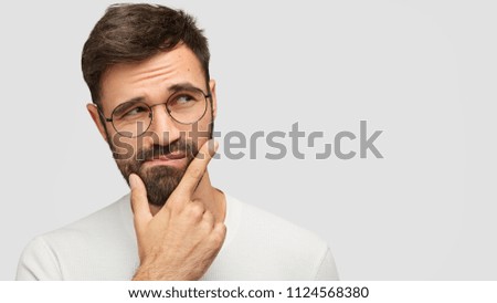Close up portrait of handsome dark haired male holds chin and looks with hesitsation aside, chooses what to buy between two things, isolaed over white background with copy space for your text