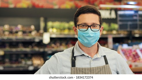 Close Up Portrait Of Handsome Joyful Male Food Store Worker In Medical Mask And Apron Looking At Camera While Standing In Grocery. Caucasian Man Indoors With With Blurred Background. Work Concept