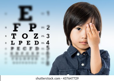 Nahaufnahme des Porträts eines gut aussehenden, kleinen asiatischen Jungen, der das Augenlicht testet. Kid, der ein Auge mit der Hand aus der scharfen Augentestkarte im Hintergrund abschließt.