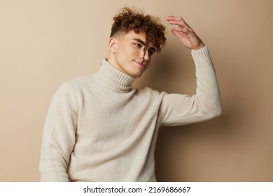 a close portrait of a handsome man with curly hair, standing on a beige background in a light turtleneck and thoughtfully holding his hand on his head touching his hair - Powered by Shutterstock