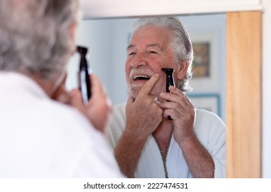 Close up portrait of a handsome senior man in bathrobe shaving beard with an electric razor looking at himself in front of the mirror - Powered by Shutterstock