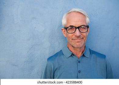 Close Up Portrait Of Handsome Senior Man With Glasses Looking At Camera, He Is Standing Against A Blue Wall.