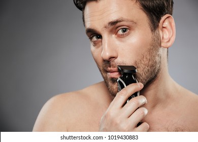 Close Up Portrait Of A Handsome Shirtless Man Shaving Beard With An Electric Razor Isolated Over Gray Background