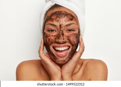 Close up portrait of happy African American woman applies coffee mask for cleansing, wants to look refreshed and healthy, poses alone against white background, shows healthy body after spa procedure