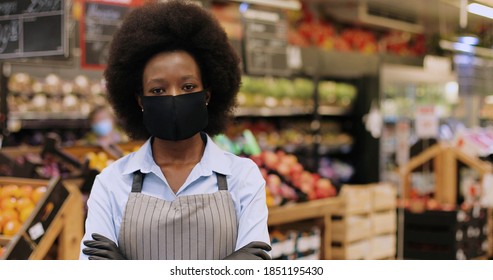 Close Up Portrait Of Happy African American Female Worker In Black Mask And Gloves Standing In Supermarket And Looking At Camera. Young Woman Food Store Assistant At Work In Quarantine