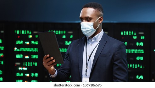 Close up portrait of happy African American man in medical mask standing among servers videochatting on tablet while checking big data processors. Male analytic speaking in video in data storage