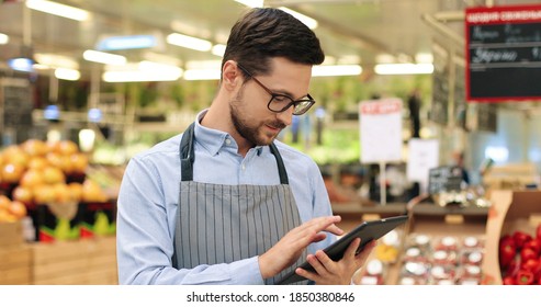 Close Up Portrait Of Happy Caucasian Male Worker In Glasses Standing In Supermarket And Typing On Tablet. Young Joyful Man Food Store Assistant At Work Tapping On Device Indoors. Retail Concept