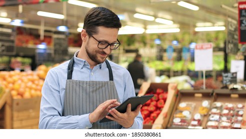 Close Up Portrait Of Happy Caucasian Male Worker In Glasses Standing In Supermarket And Typing On Tablet. Young Joyful Man Food Store Assistant At Work Tapping On Device Indoors. Retail Concept