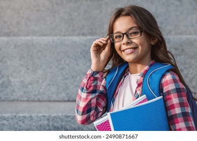 Close up portrait of happy caucasian young preteen girl elementary middle school pupil in eyeglasses with backpack holding notebook books outside the primary school. Education concept copy space - Powered by Shutterstock