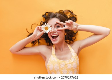 Close up portrait of happy girl showing victory sign by face lying on yellow floor. Brown-haired woman in T-shirt smiles broadly with her teeth. Good mood concept, leisure