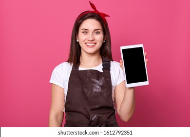 Close up portrait of happy housewife dresses dirty apron, beautiful girl looking at camera with charming smile and holds white tablet with blank sreen. Copy space for advertisment or promotional text. - Powered by Shutterstock