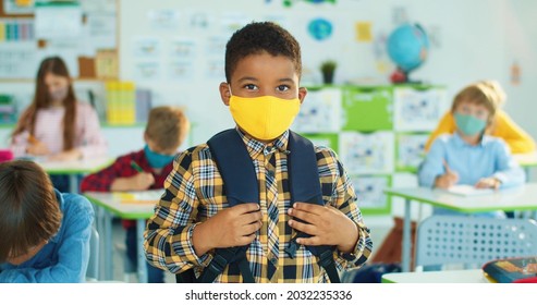 Close up portrait of happy little cute African American boy in yellow face mask looking at camera while standing in classroom. Elementary school, primary education. Coronavirus pandemic, new normal - Powered by Shutterstock