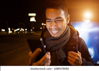 Close up portrait of happy middle aged asian man looking at his mobile phone and smiling outdoors in city at night - Powered by Shutterstock