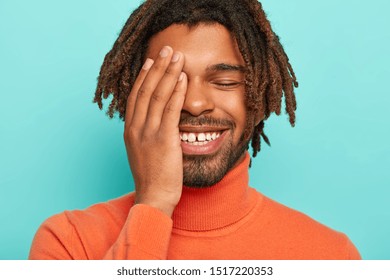 Close up portrait of happy pleased African American man smiles broadly, has white teeth with little gap, covers face with palm laughs at funny situation happened with him poses indoor wears turtleneck
