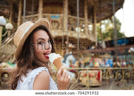 Close up portrait of a happy pretty girl eating ice cream while standing in front of the carousel at the amusement park