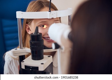 Close up portrait of happy smiling female child sitting in front of the experienced doctor while checking eyesight with slit lamp in ophthalmology cabinet