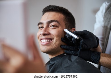 Close Up Portrait Of Happy Smiling Indian Man In Black Peignoir Looking Away While Barber Cutting His Hair With Electrical Tool In Barber Shop