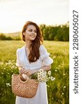 close portrait of a happy woman looking at the camera in a light dress and a wicker basket in her hands with chamomile flowers in nature