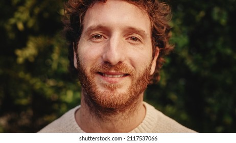 Close Up Portrait Of A Happy Young Adult Male With, Brown Eyes, Curly Ginger Hair And Beard Posing For Camera. Handsome Diverse Caucasian Male Smiling On Green Nature Background.