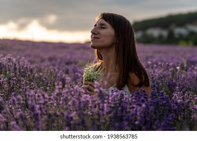 Close up portrait of happy young brunette woman in white dress on blooming fragrant lavender fields with endless rows. Warm sunset light. Bushes of lavender purple aromatic flowers on lavender fields.
