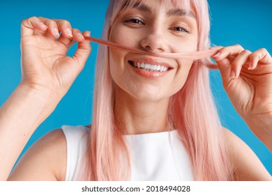 Close up portrait of happy young woman with natural long pink dyed hair holding a strand of hair as a moustache and smiling at camera, posing isolated over blue studio background. Hair care concept