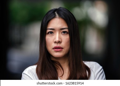 Close Up Portrait Head Shot Of The Face Of A Beautiful, Young And Attractive Thai Chinese Asian Woman In A Casual White Shirt Glaring Into The Camera. She Looks Serious, Concerned Or Worried.