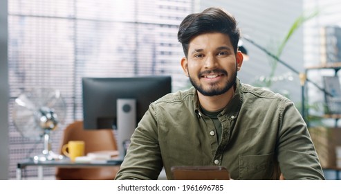 Close Up Portrait Of Hindu Joyful Young Handsome Male Worker Texting On Tablet Browsing Online While Sitting At Workplace In Cabinet Looking At Camera And Smiling Office Concept Successful Businessman