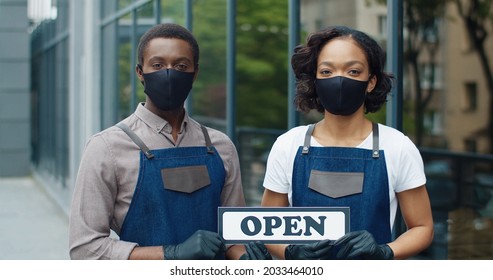 Close Up Portrait Of Joyful African American Young Man Woman Couple In Apron And Black Mask Standing Outdoors Near Coffee Shop With Open Sign And Looking At Camera. Restaurant Business, Quarantine