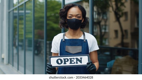 Close Up Portrait Of Joyful Beautiful African American Young Woman In Apron And Black Mask Standing Outdoor On Street With Open Sign And Looking At Camera Restaurant Business, Coffee House, Quarantine