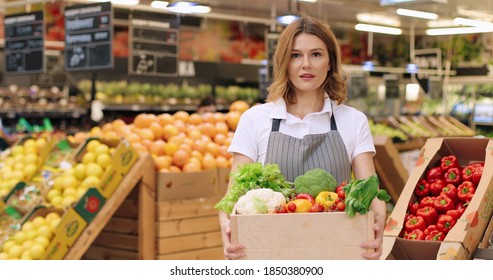 Close Up Portrait Of Joyful Caucasian Female Supermarket Worker In Good Mood Holding In Hands Box With Fresh Vegetables, Looking At Camera And Smiling. Young Woman Seller With Organic Food Job Concept