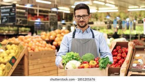 Close Up Portrait Of Joyful Caucasian Male Supermarket Worker In Good Mood Holding In Hands Box With Fresh Organic Vegetables, Looking At Camera And Smiling. Young Man In Glasses With Organic Food