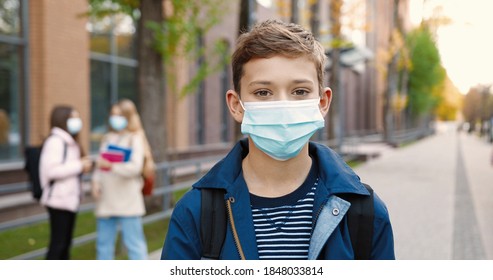 Close up portrait of joyful cute schoolboy standing outdoors with backpack and putting off medical mask. Male Caucasian student in mask near school smiling to camera. Quarantine concept