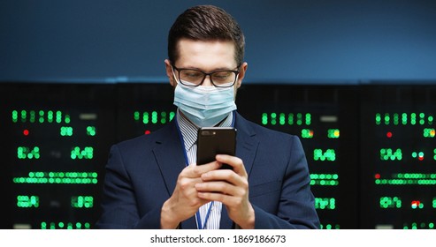 Close up portrait of joyful handsome Caucasian male businessman in mask standing in server room with many processors and texting on cellphone at work. Man worker typing on smartphone. Tech concept