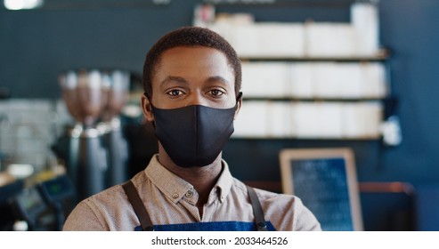 Close Up Portrait Of Joyful Handsome Young African American Male Waiter In Black Mask Standing In Cafe Looking At Camera In Good Mood. Man Worker In Coffee Shop Indoor At Workplace. Job Concept