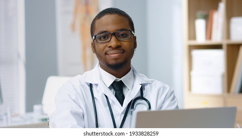 Close Up Portrait Of Joyful Happy Young African American Male Physician In Glasses Sitting In Hospital Cabinet Alone, Looking At Camera And Smiling. Handsome Doctor In White Coat With Smile On Face