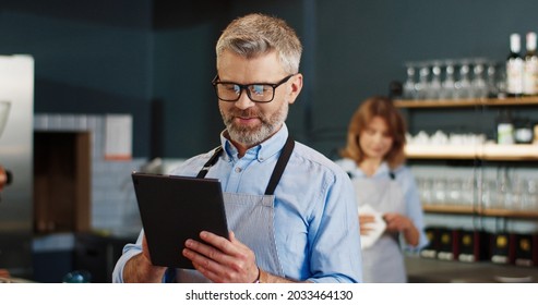 Close up portrait of joyful middle-aged bearded businessman in apron tapping on tablet device browsing online standing in restaurant. Coffee shop concept, coffee house, family business - Powered by Shutterstock