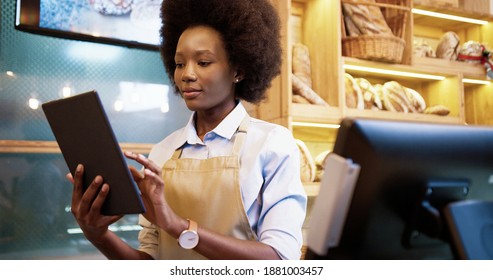 Close up portrait of joyful young African American beautiful female seller worker standing in small bakehouse in good mood typing and scrolling on tablet at workplace. Bakery worker concept