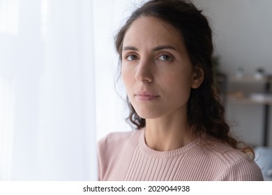 Close up portrait of joyless 30s woman standing alone near window indoor. Face of Hispanic serious female with sad eyes staring at camera. Tiredness, lack of optimism, solitude, life concerns concept - Shutterstock ID 2029044938
