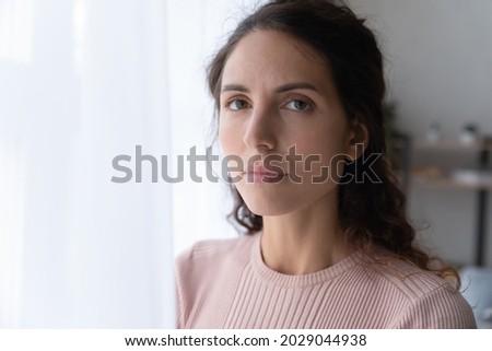 Close up portrait of joyless 30s woman standing alone near window indoor. Face of Hispanic serious female with sad eyes staring at camera. Tiredness, lack of optimism, solitude, life concerns concept