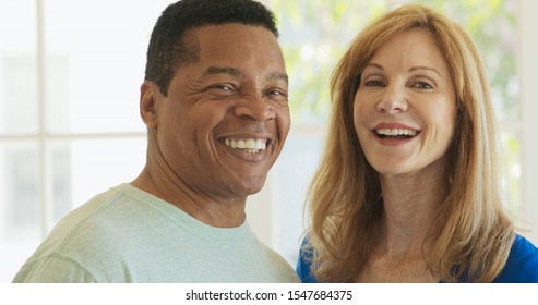 Close Up Portrait Of Laughing Mixed Race Senior Couple Looking At Each Other Then Turning To Camera. Happy Older African American Husband And Caucasian Wife At Home