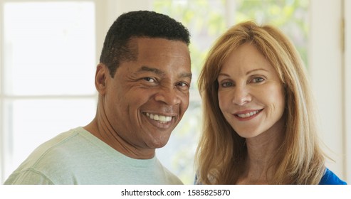 Close Up Portrait Of Laughing Mixed Race Senior Couple Looking At Each Other Then Turning To Camera. Happy Older African American Husband And Caucasian Wife At Home