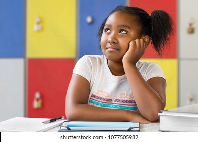 Close Up Portrait Of Little African Female Student Sitting At Desk In Classroom. Tedious Kid With Bored Wondering Facial Expression Staring And Looking Up.