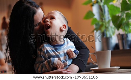 Close up portrait of a little boy with special needs and mom laughing at a table in a cafe, lifestyle. Mom's love for her child, inclusion.Happy disability kid concept.