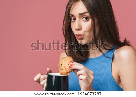 Close up of portrait of a lovely young attractive brunette woman standing isolated over pink background, holding a cup of coffee and eating a chocolate chip cookie